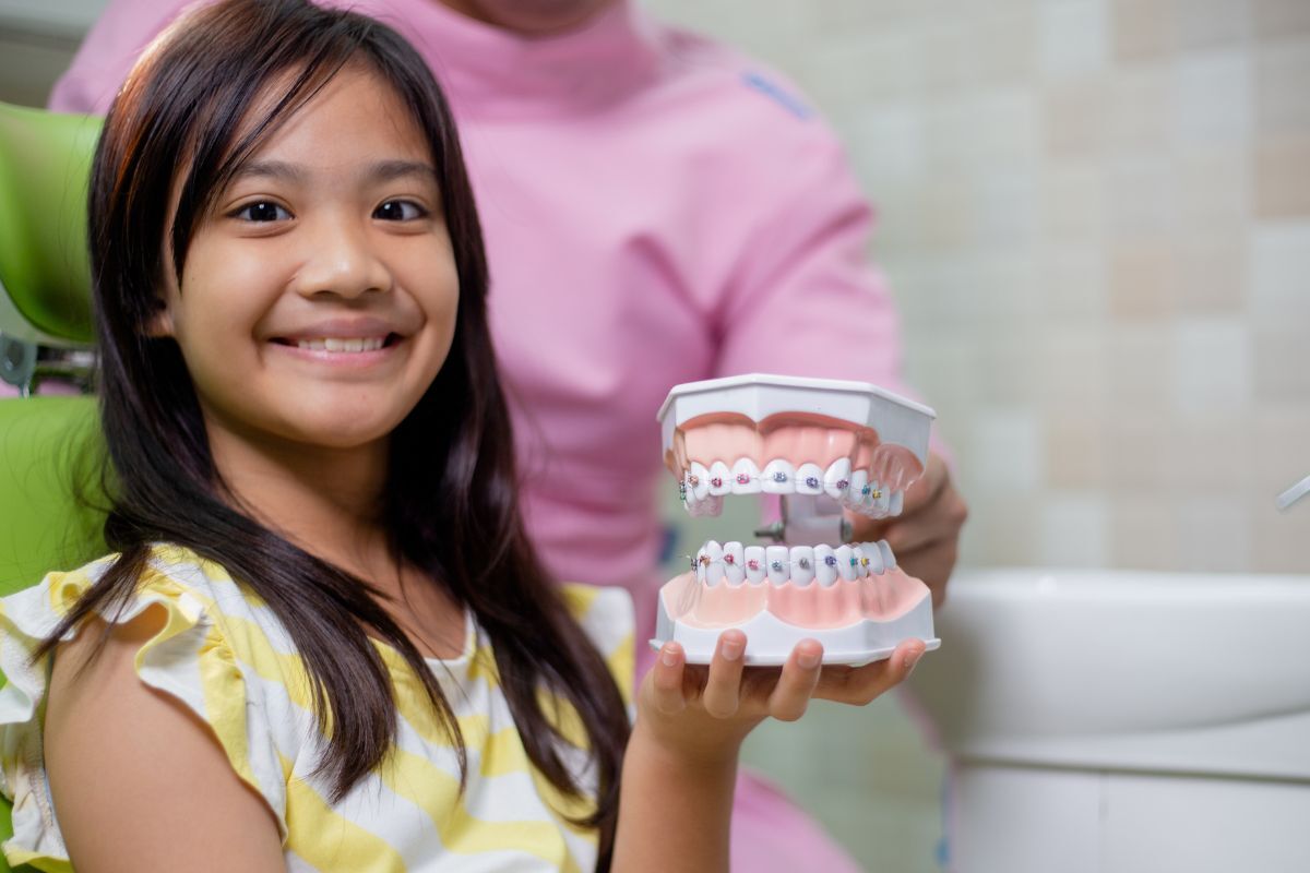 Young girl smiling while holding orthodontic model showing braces treatment for bite correction.