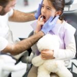 Pediatric dentist examining child's teeth in dental chair with stuffed animal for comfort.
