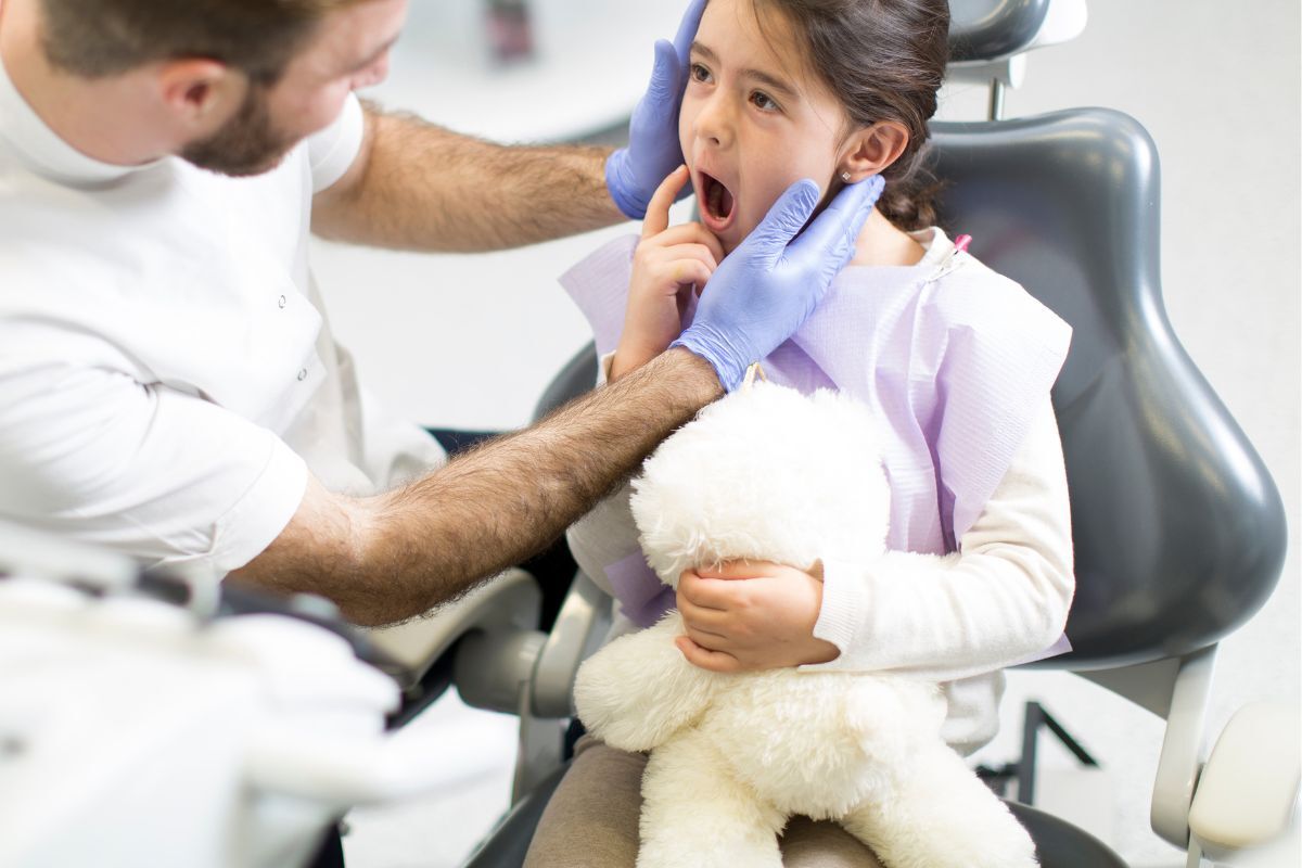 Pediatric dentist examining child's teeth in dental chair with stuffed animal for comfort.