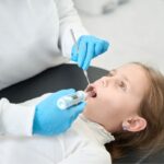 Pediatric dentist examining child's teeth with dental mirror during oral sedation consultation.