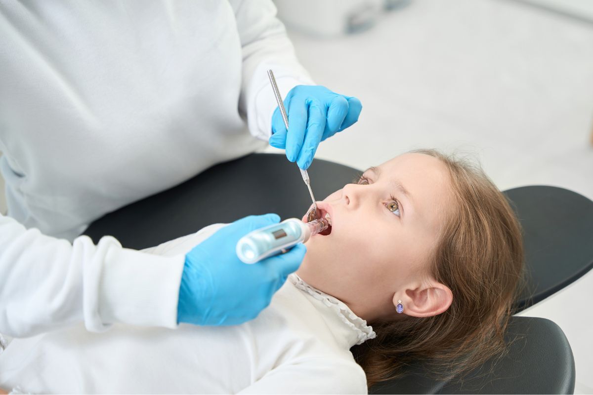 Pediatric dentist examining child's teeth with dental mirror during oral sedation consultation.
