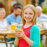 Happy girl holding juice glasses at outdoor family gathering, promoting healthy drink choices.