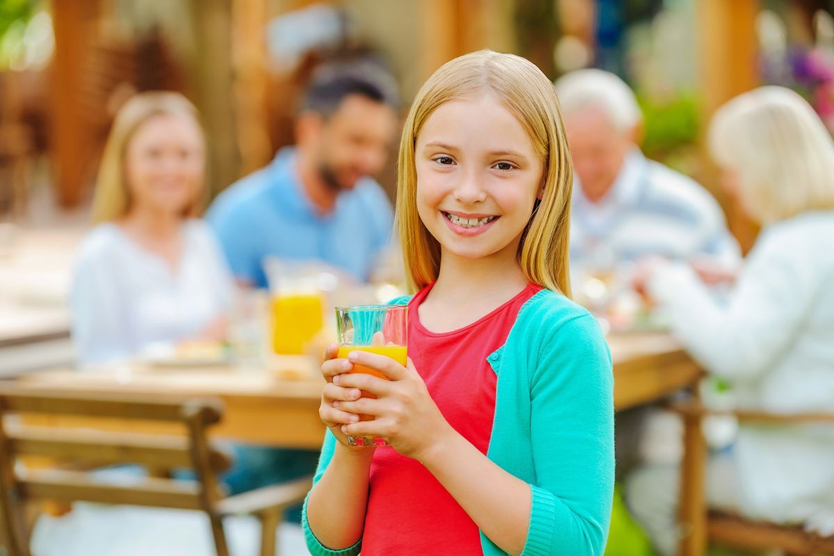 Happy girl holding juice glasses at outdoor family gathering, promoting healthy drink choices.
