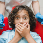Nervous child covering mouth with hands during dental visit with gloved dentist.