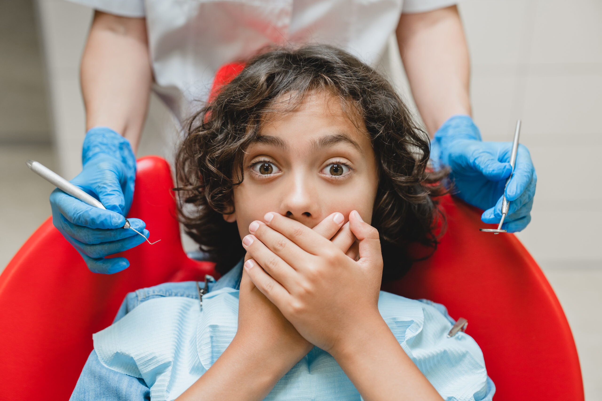 Nervous child covering mouth with hands during dental visit with gloved dentist.