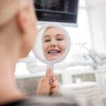 Young patient with metal braces smiling at mirror in dental office.
