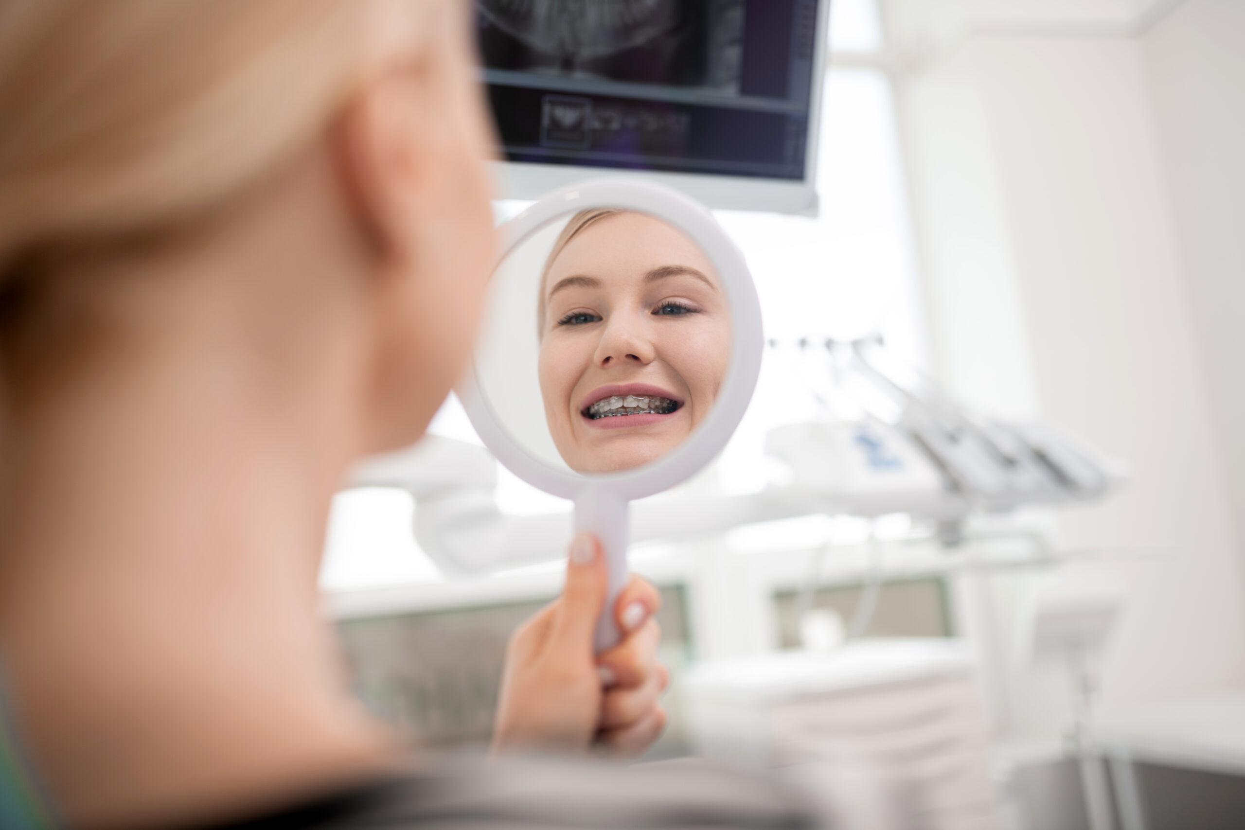 Young patient with metal braces smiling at mirror in dental office.