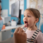 Young girl with open mouth during pediatric dental exam with tongue depressor.