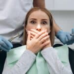 Anxious patient covering mouth during dental examination with dentist holding tools.