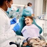 Young boy smiles in dental chair while pediatric dentist prepares for examination in Spring.